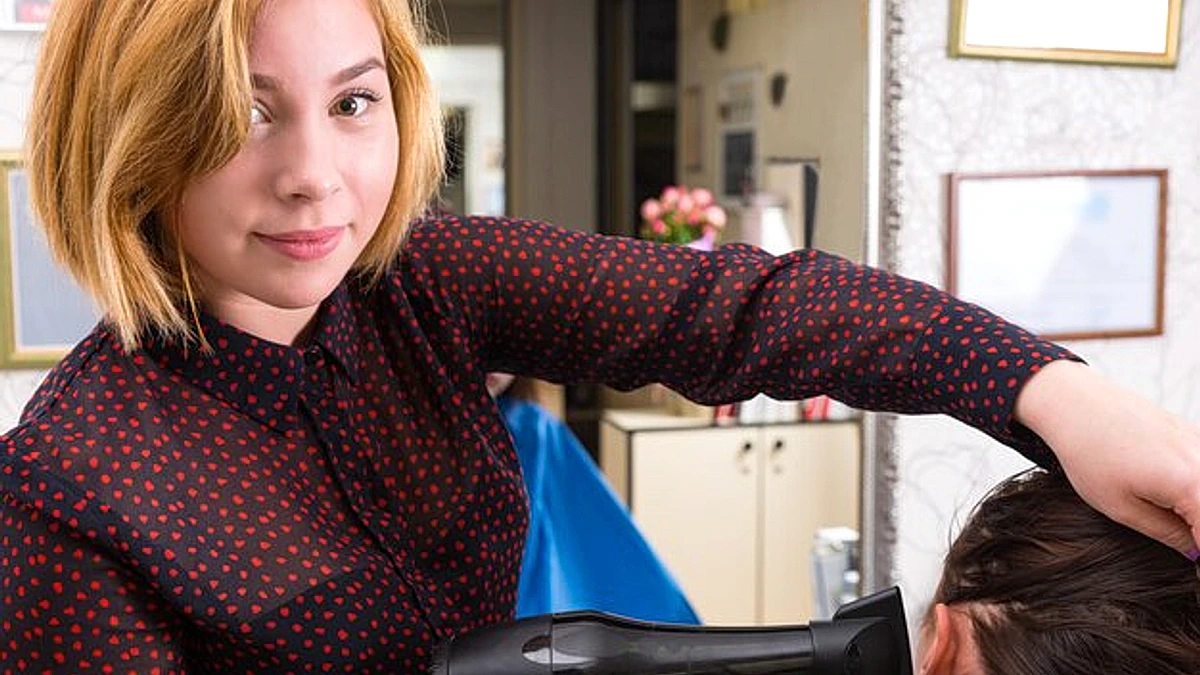 Close Up Portrait of Young Blond Female Stylist Drying Hair of Brunette Woman with Blow Dryer in Salon with Large Mirror in Background